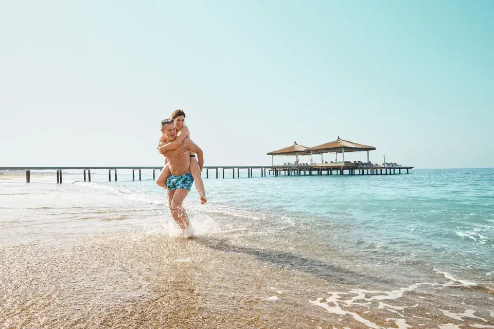 Das Bild zeigt ein Paar am Strand auf den Malediven mit einem Steg im Hintergrund, dabei trägt der Mann seine Partnerin Huckepack.