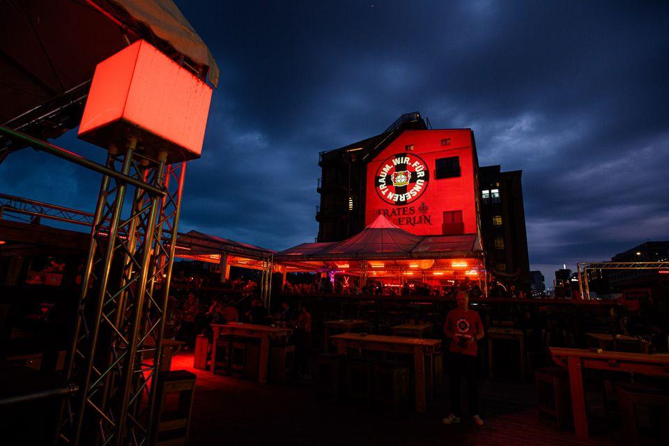 Das Bild zeigt ein Event in einer Aussenlocation mit vielen Menschen und Ständen und dem Logo von Bayer 04 Leverkusen auf ein Gebäude projiziert.